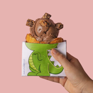 Mano sosteniendo un Dinocono de waffle con helado de chocoavellana sobre un fondo rosa pastel.